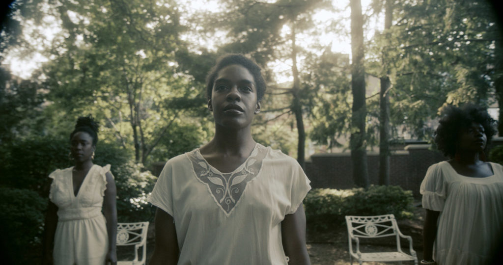 three Black women in white dresses stand in a sun-spotted green area, surrounded by tall trees and two white vintage chairs